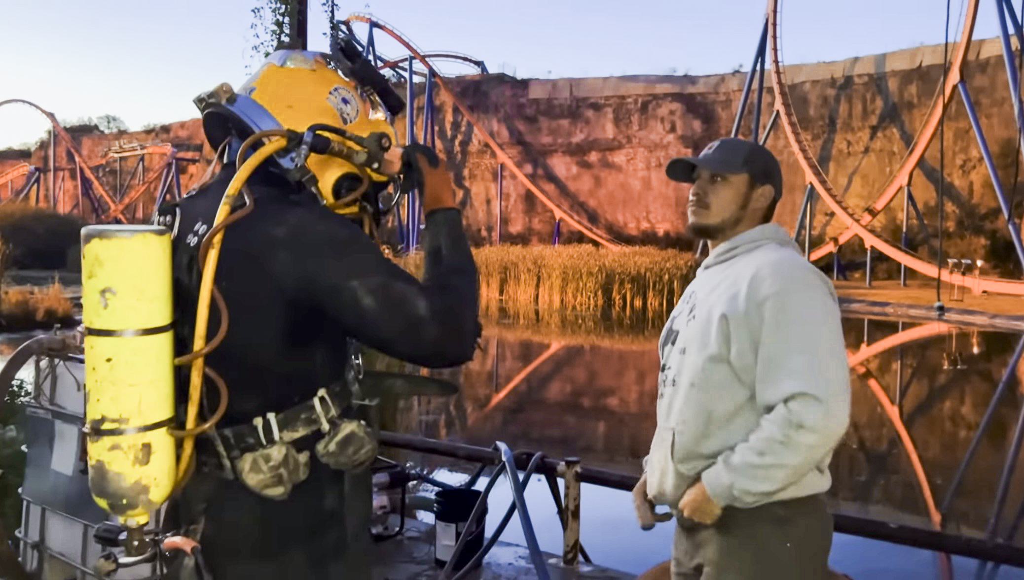 A commercial diver in a black drysuit and yellow SL-27 diving helmet stands on a pier discussing project details with a teammate at the Six Flags park pond, with a rollercoaster in the background.